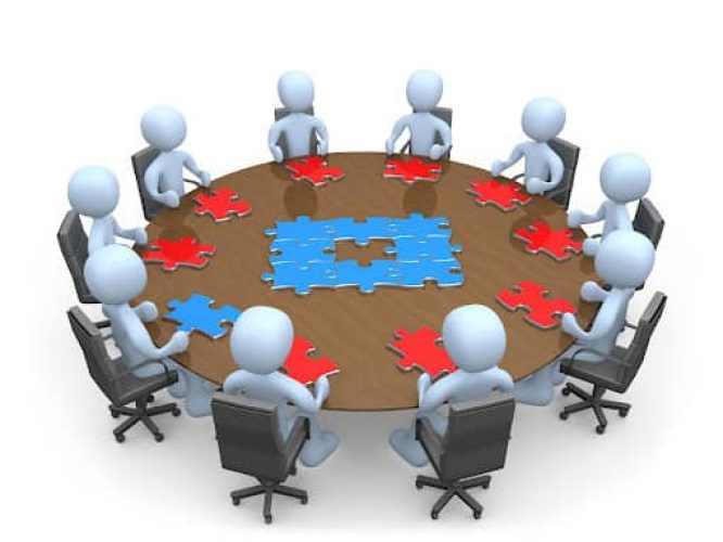 Royalty-free 3d business computer generated clipart graphic picture of a group of light blue people holding a meeting and trying to solve a jigsaw around a large rectangular conference table in an office. All but one of them have red puzzle pieces. One person holds the final blue piece to the puzzle.
